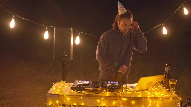 Medium shot of energetic male dj in headphones adjusting sound and mixing music on deck decorated with string lights at open-air party at night