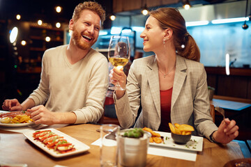 Smiling cheerful couple sitting in a restaurant, having dinner and chatting. Man talking to a woman while a woman listening to him and drinking white wine. - 391670272