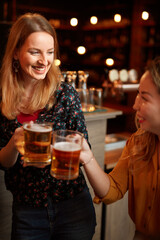 Two female friends standing in a pub and toasting with fresh cold beer.