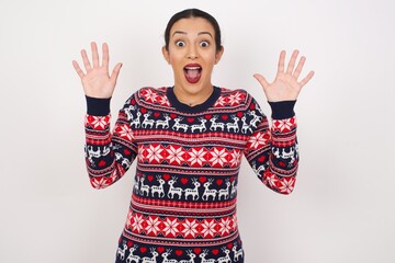 Optimistic Young beautiful Arab woman wearing Christmas sweater, against white background raises palms from joy, happy to receive awesome present from someone, shouts loudly, Excited model screaming.