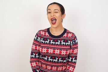 Young beautiful Arab woman wearing Christmas sweater, against white background winking looking at...