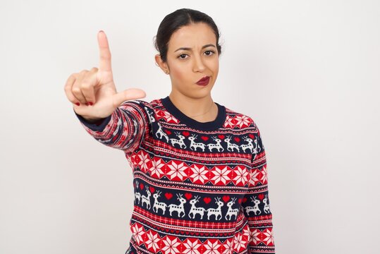 Young beautiful Arab woman wearing Christmas sweater, against white background making fun of people with fingers on forehead doing loser gesture mocking and insulting.