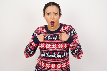 Embarrassed Young beautiful Arab woman wearing Christmas sweater, indicates at herself with puzzled expression, being shocked to be chosen to participate in competition, hesitates about something