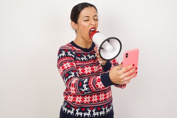 Crazy impressed Young beautiful Arab woman wearing Christmas sweater against white wall,  use cell...