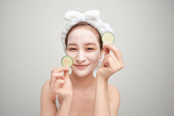 Beautiful young woman with facial mask on her face holding slices of fresh cucumber.