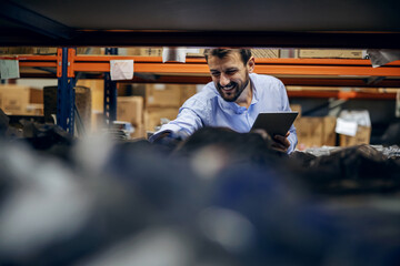 Smiling bearded boss standing in warehouse, holding tablet and checking on supplies on shelf.