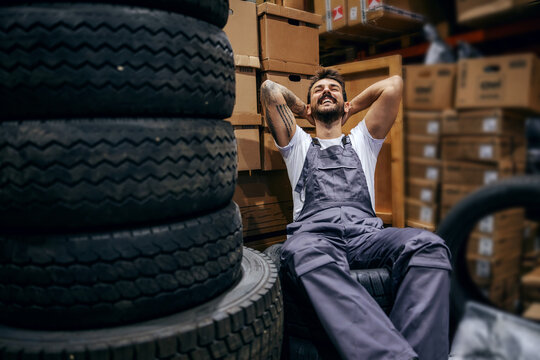 Smiling bearded tattooed worker sitting on tires in storage of import and export firm. He is taking a break from hard work.