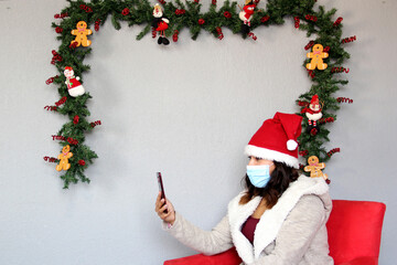 
Latin woman with protection mask and Santa Claus hat on video call with cell phone and laptop....