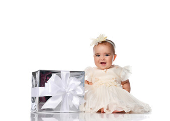 Little baby princess in a smart white dress sits in a white studio with a gift box with a ribbon.