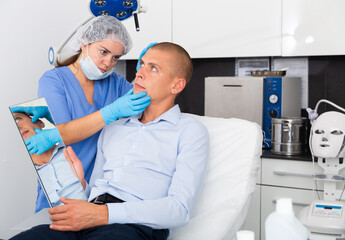 Confident woman cosmetologist examining male client face before procedure in esthetic clinic. Man looking at mirror