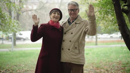 Happy elegant senior couple looking at camera and waving. Portrait of cheerful smiling Caucasian husband and wife posing outdoors on autumn day in city park. Family leisure and happiness concept.