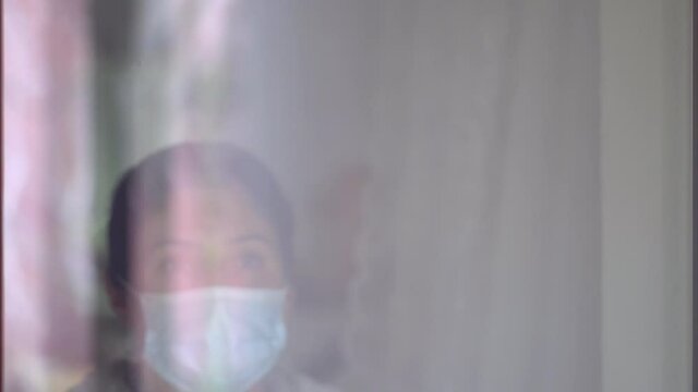 Young caucasian woman in a coronavirus mask touching with her hand to the glass of the window. Sad woman in quarantine. Pensive young woman in mask to protect herself from covid-19 while looking out