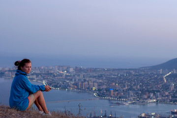 Fototapeta na wymiar a girl in a blue sweater sits on a mountain overlooking the city, the concept of hiking in the mountains, picnic, tracking. looks to the side