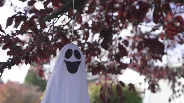 Close up tracking shot of a creepy Halloween ghost decoration hanging on a blossom tree in Autumn and swaying with the wind.