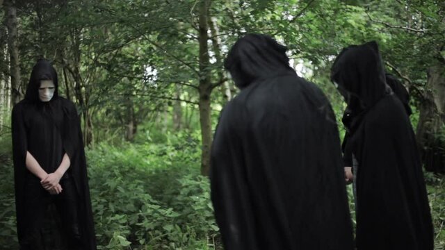 A Group Of Pagan Cult Members Gather In A Ritualistic Circle In The Woods. The Creepy Group Of Hooded And Masked Figures Are Holding Staffs And Fire Torches.