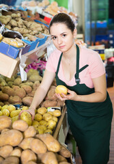 young female grocery worker in apron arranging vegetables on shop shelves