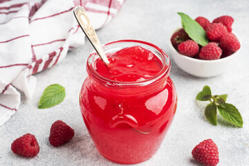 Homemade raspberry jam in a glass jar and fresh raspberries with mint on a grey concrete...