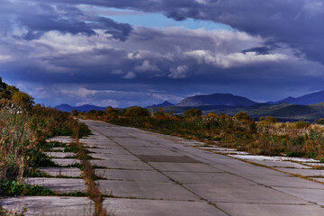 Abandoned concrete runway for military aircraft from Russia. Against the backdrop of the runway wild field, mountains and gloomy sunset clouds. Stylish picture from the hinterland of Russia.