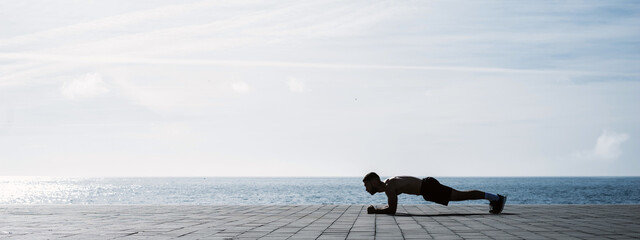 Fit young man doing plank on seafront. Fitness training in morning outdoors. Working out during...