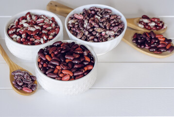 red and colored beans in white bowls and wooden spoons on a white background. background with different types of beans.
