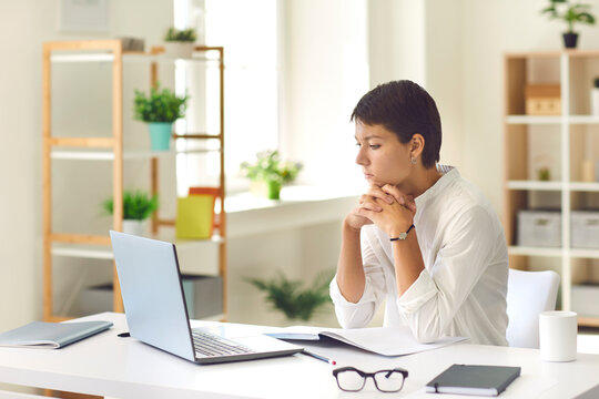 Pensive woman sitting at desk with laptop and notebook watching educational video or reading ebook