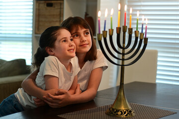 Two happy Jewish sisters looking at a beautiful menorah candelabra glowing on the eight day of...