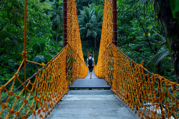 Hanging bridge with woman tourist and orange ropes in green jungle forest. Sportive girl trekking alone in jungle forest. Tropical nature landscape. Suspension bridge view. Rope bridge river crossing