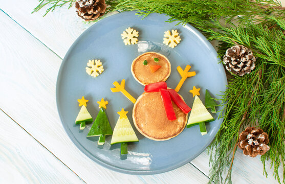 Christmas pancake shaped like a snowman with fresh vegetables blue plate on wooden white table decorated festively. Christmas fun food for kids, funny breakfast brunch idea xmas
