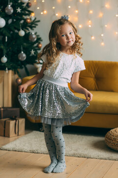 A Happy Little Girl With Blonde Hair In An Elegant Sequined Skirt Is Standing In The Living Room Next To A Decorated Christmas Tree. Christmas