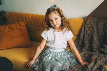A happy little girl with blonde hair in a shiny skirt is sitting on a yellow sofa in the living room.
