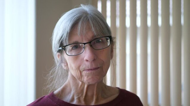 A happy old caucasian woman with aging face wrinkles gray hair and glasses looking at camera with a sad but hopeful outlook for her life.