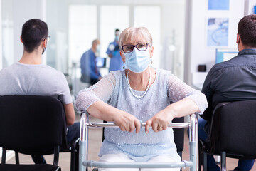 Disabled senior woman with face mask against coronavirus and walking frame looking at camera in...