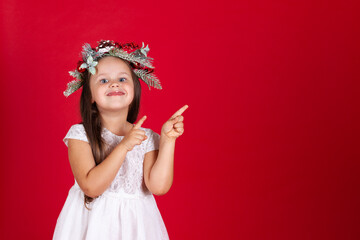 close up of a smiling girl in a Christmas wreath pointing with her index fingers at an empty space for text on a red background.