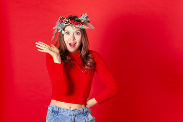 close up portrait of a surprised and dissatisfied young woman in a Christmas wreath, hands on hips, isolated on a red background.