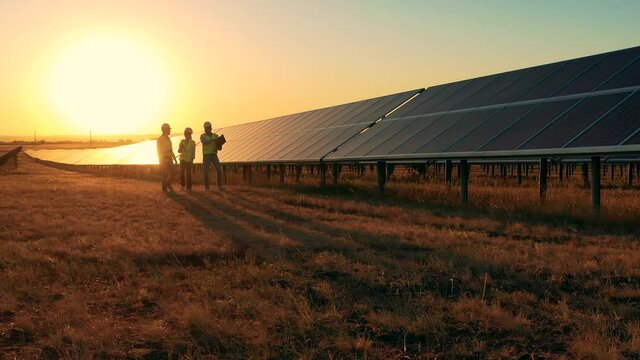 Three solar energy specialists walking through a solar park at sunset. Drone shot