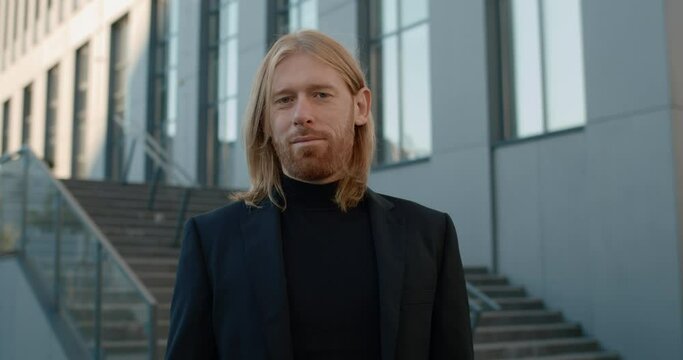 Portrait of handsome man looking to camera while using smartphone. Crop view of successful businessman with beard and long hair smiling while standing at stairs outdoors. Zoom in.