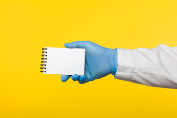 Close-up profile side view of human hand in blue surgical gloves holding notebook in hand and holding blank paper. On a yellow background.