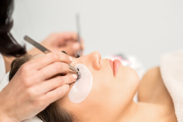 Young caucasian woman having eyelash extension procedure in beauty salon. Beautician glues eyelashes with tweezers