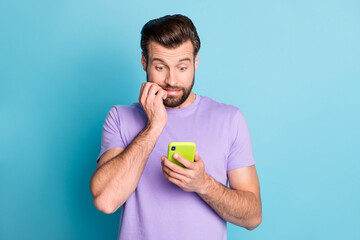 Photo portrait of astonished man keeping smartphone biting fingers staring problem isolated vivid...