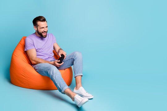 Full length body size photo of millennial playing console controller sitting in beanbag isolated on vivid blue color background empty space