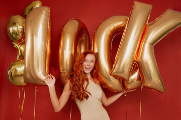 Inspired european long-haired woman dressed shine dress and holding balloons. Winsome red haired girl posing on red background.