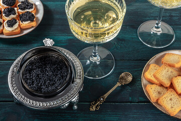 Caviar in a vintage bowl with a champagne coupe glass, with bread and toasts, on a dark blue wooden background - obrazy, fototapety, plakaty
