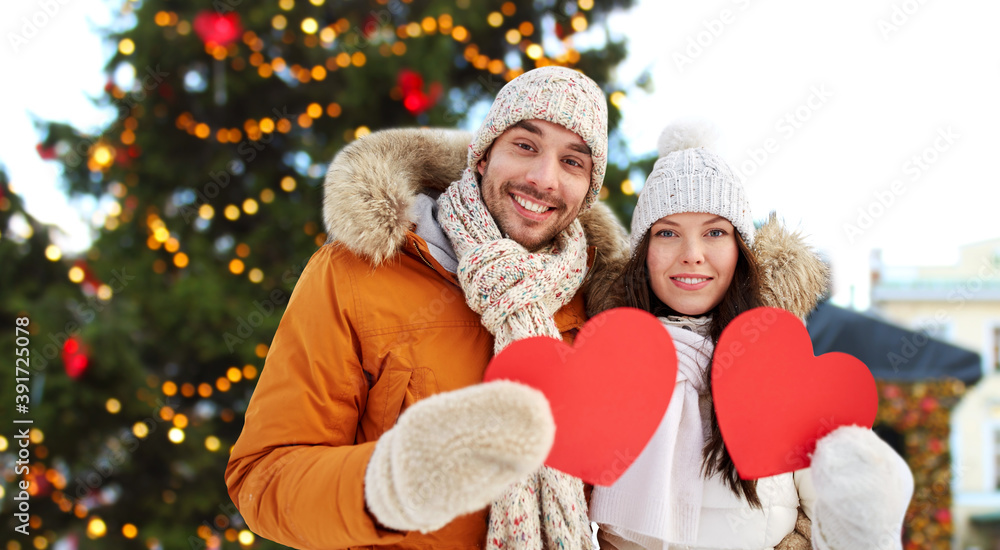 Wall mural winter holidays, love and people concept - happy couple holding blank red hearts over christmas market background