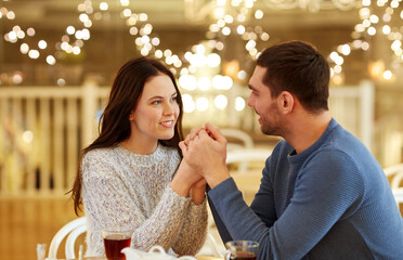 love, romance and dating concept - happy couple drinking tea and holding hands at cafe or restaurant over festive lights