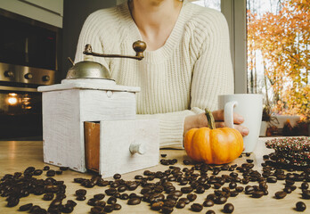 Young girl in warm, cosy knitted autumn sweater, leaning on a kitchen counter with vintage retro coffee grinder, small pumpkin, cookies and a cup. Unground coffee beans scattered on the table.