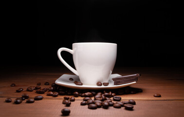 Small cup of hot coffee, pieces of delicious chocolate bar and fresh unground coffee beans scattered on a wooden table, with black background.