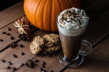 Tall glass of delicious coffee with whipped cream and chocolate sprinkles. Autumn composition on a wooden table, with cinnamon sticks, star anise, pumpkin, chocolate chip cookies and coffee beans.