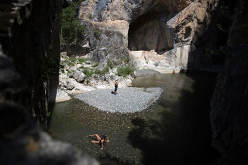 Photo from a stone bridge,of a woman who lie down inside the cold water of Venetikos river.A man admires the high cliffs of 200 meters of the magnificent Gorge.
