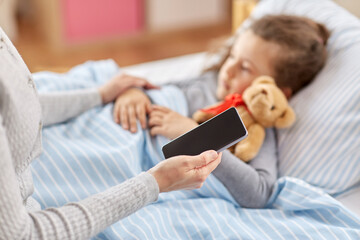family, health and people concept - sick little daughter sleeping in bed and mother with smartphone...