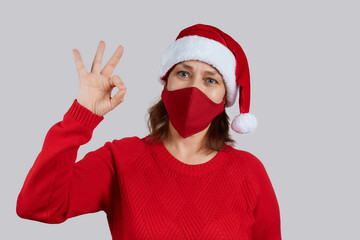 Fototapeta na wymiar An adult woman in a red protective mask, in a Santa hat on a gray background shows the gesture ok. Christmas, New Year in quarantine